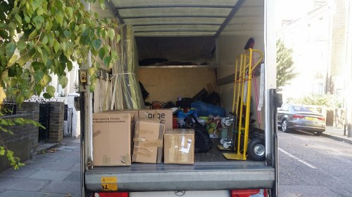 The image shows the open rear of a delivery van parked on a pavement in a residential area, with a variety of waste materials visible inside. In the foreground, several large cardboard boxes with printed labels and logos are stacked, some appearing to contain other items, while others are flattened or damaged. Behind the boxes, there is an assortment of plastic bags, loose packaging materials, and miscellaneous rubbish, including some plastic containers and household waste. To the right side of the van, a yellow hand truck or dolly is leaning against the vehicle, and a small vacuum cleaner or similar device is placed on the pavement nearby. The background reveals a typical street scene with parked cars, green trees, and residential buildings, suggesting a local Southgate or North London environment. The scene likely depicts a rubbish removal process undertaken by a service such as Commercial Waste Southgate, emphasizing their capability to clear bulky or household waste from property driveways or commercial premises.