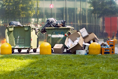 Two large industrial wheelie bins are positioned on a paved area in front of a modern glass building, with the left bin being green and the right bin also green. Both bins have black plastic bags partially covering their open tops, with some bags hanging over the sides. To the right of the green bins, there is a noticeable pile of mixed rubbish, including flattened cardboard boxes, some slightly torn, and scattered smaller packaging materials. An orange plastic crate filled with various waste is visible beside the cardboard, along with additional loose cardboard and plastic items. The rubbish is arranged on a paved surface, bordered by a strip of green grass in the foreground, and yellow bollards are situated along the edge of the pavement, acting as barriers. The environment appears to be an outdoor commercial or industrial area, possibly adjacent to a business premises in Southgate, with reflections of trees and nearby structures visible in the glass facade behind the waste collection. The overall scene suggests a waste collection or rubbish removal activity undertaken by Commercial Waste Southgate in the local area. The lighting indicates daytime with natural sunlight illuminating the scene clearly.