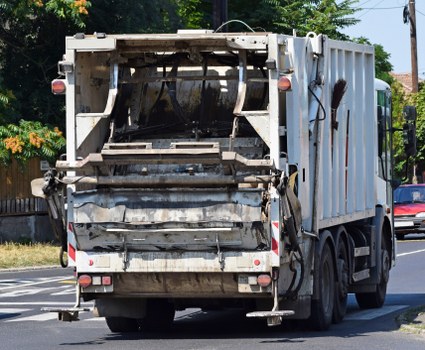 A large commercial waste collection truck, primarily white with some visible dirt and grime, is positioned on a city street, likely in Southgate South London. The rear-loading mechanism is extended and appears to be designed for compacting and lifting rubbish, with signs of wear and use evident in the metal finish and hydraulic components. The truck's rear doors are closed, with a small section open to accommodate waste collection, and the textured metal surface shows areas of dirt accumulation. Visible safety markings, including reflective red and white stripes, are present along the lower edge of the truck. In the background, a sidewalk, trees with green foliage, and a few additional vehicles, including a red car, are visible, indicating the urban environment. Overhead power lines and street signage suggest a typical residential or commercial street setting. The scene portrays everyday rubbish removal activity undertaken by Commercial Waste Southgate, capturing the operational aspect of business waste collection in the area, with natural daylight highlighting the truck's utilitarian finish and the surrounding environment.