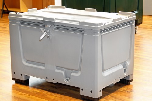 A large, beige plastic waste container with a hinged, tightly fitting lid secured by a metal clasp, placed on a polished wooden floor in an indoor environment. The container has a textured surface with subtle horizontal ridges and is supported by four black rubber feet at the base. Behind it, there are green and brown waste bins, indicating a setting related to rubbish management, possibly within a commercial or communal space. The container is designed for heavy-duty use, suitable for business waste removal services, such as those provided by Commercial Waste Southgate, and is positioned in an organized manner for waste collection or disposal purposes.