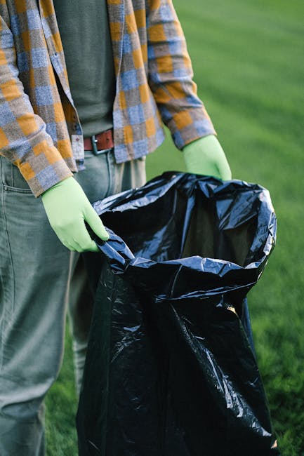 Close-up of a person wearing a plaid shirt, green gloves, and grey trousers, holding open a large black rubbish bag with both hands, positioned outdoors on a grassy area. The bag is made of shiny, dur