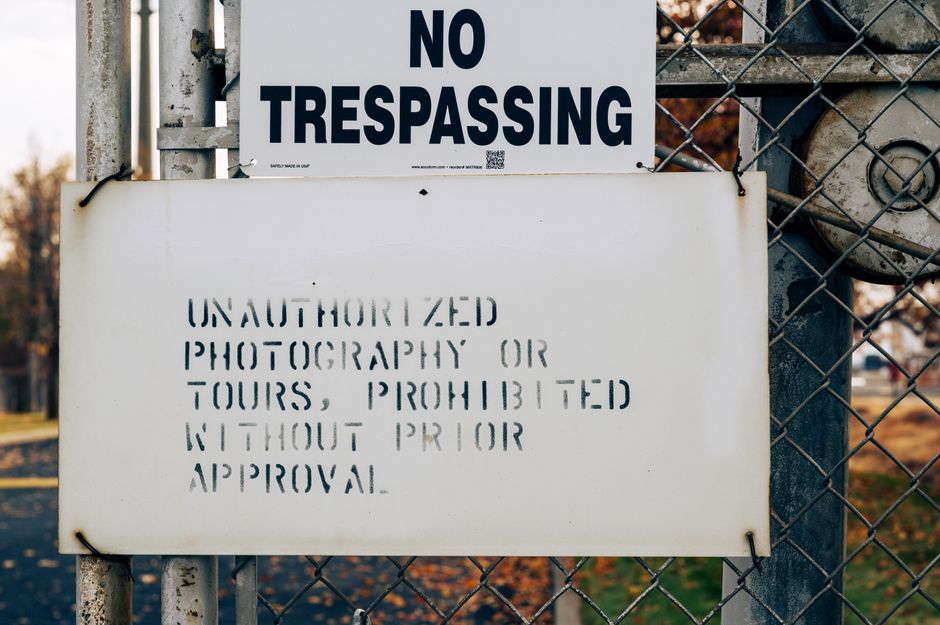 Close-up of a metal gate with two signs attached. The upper sign reads 'NO TRESPASSING' in bold black letters on a white background. The lower sign, also on a white background, displays the message 'U
